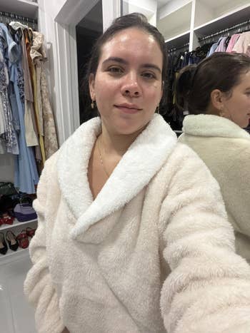 Person in a plush, cozy robe stands in a walk-in closet with clothing and shoes visible in the background