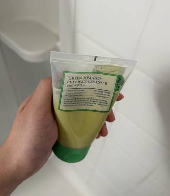 Person holding a tube of Green Tomato Clay Pack Cleanser in a bathroom