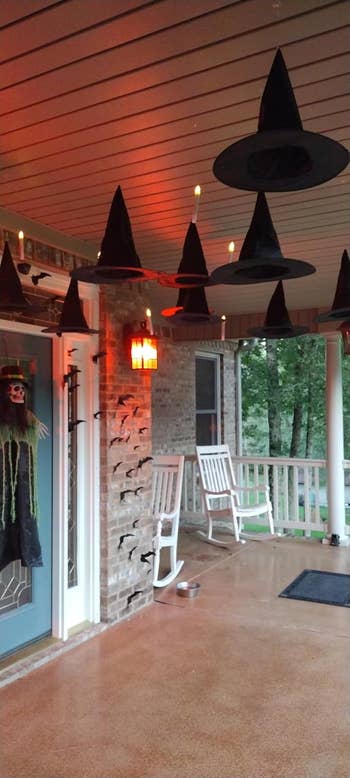 Witches' hats with lights hang from a porch ceiling, next to Halloween decorations like a ghoul figure on a door. Rocking chairs sit nearby