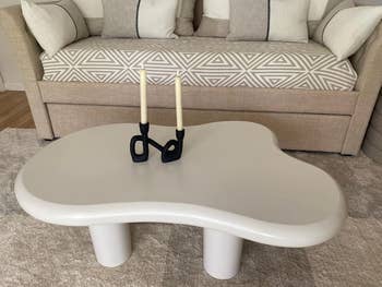 A stylish beige couch with geometric-patterned cushions and a wavy, modernist white coffee table with two black candleholders holding white candles on it