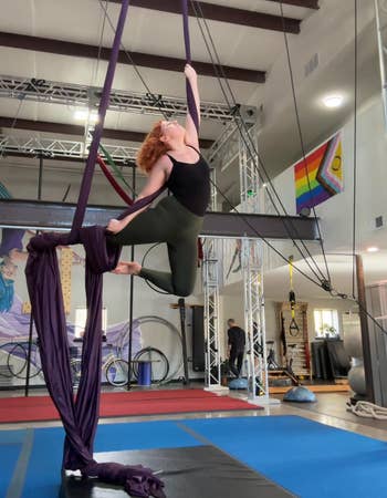 Person performing aerial silks routine in a gym, showcasing athletic strength and flexibility. Fitness equipment visible in the background