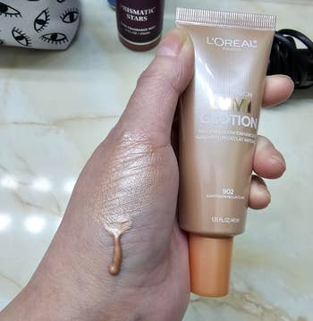 Hand holding L'Oréal liquid foundation tube with product dribbling onto skin, conveying texture and consistency for shopping consideration