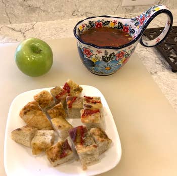 A decorative mug of tomato soup with a handle rests on a counter, next to a green apple and a plate of cubed bread