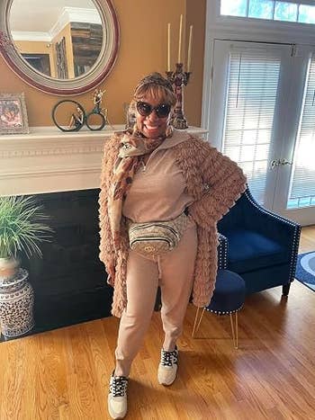 Person in a cozy textured coat, sunglasses, and sneakers stands in a living room, accessorized with a patterned scarf and a small waist bag