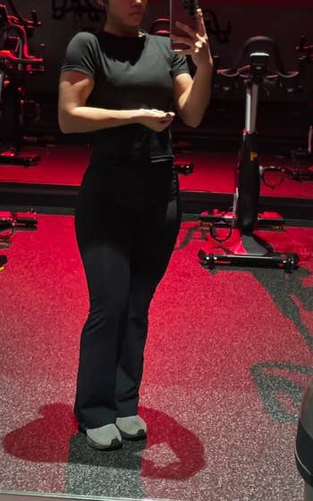 Person taking a gym mirror selfie in an exercise room with stationary bikes, wearing workout attire including a fitted T-shirt and pants