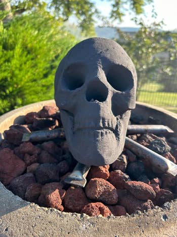 A decorative skull is placed on top of rocks and charcoals in an outdoor setting