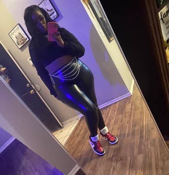 Person taking a mirror selfie, wearing a black crop top, shiny leggings, and sneakers with colorful accents, accessorized with a chain belt