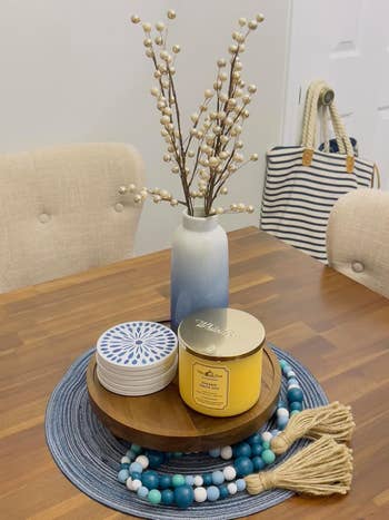 A decorated table with a vase of branches, yellow candle, blue and white coasters, and a blue beaded tassel. A striped bag hangs on a door handle in the background