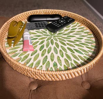 A decorative round tray holds a hair clip, a tube of pink cream, and two black remote controls on a cushioned surface