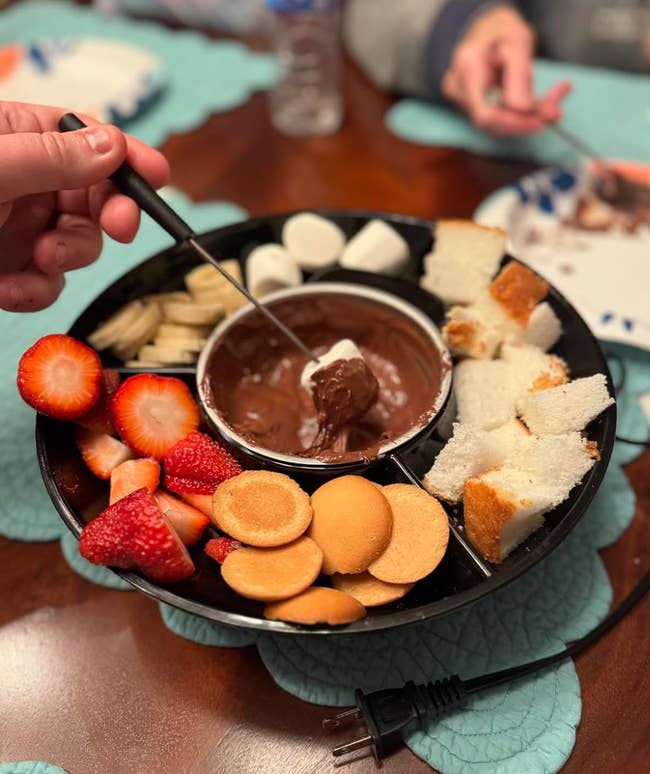 A tray of assorted snacks (strawberries, cookies, marshmallows, and cake pieces) surrounding a bowl of chocolate fondue