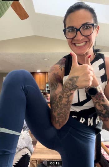 Woman in sleeveless top and leggings giving thumbs up, showing tattooed arms, indoors. No identifiable persons