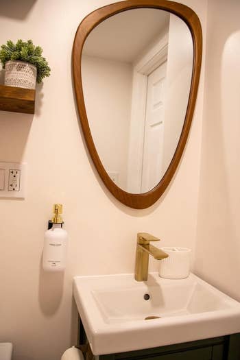 Modern bathroom showing a white sink with a gold faucet, round edge mirror, soap dispenser, and small plant on a shelf