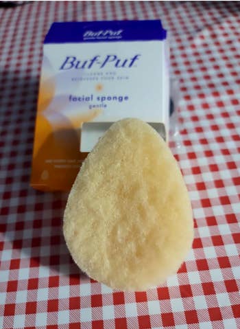 Buff Puf gentle facial sponge displayed on red checkered surface, with packaging in the background