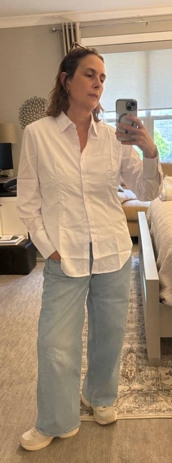 Person taking a selfie in a bedroom mirror, wearing a white button-up shirt, light jeans, and sneakers