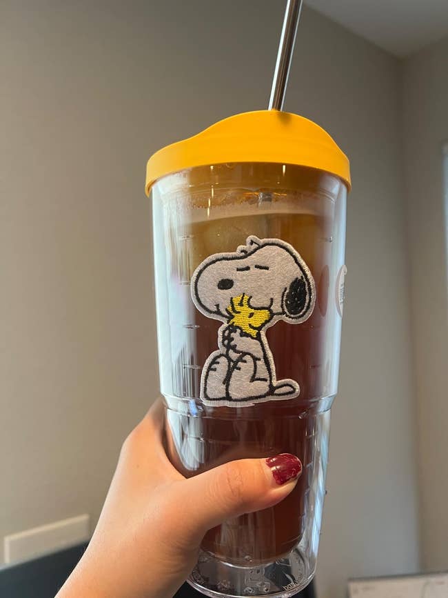Tumbler with iced drink featuring Snoopy hugging Woodstock design. Woman holding it with red polished nails