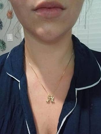 Person wearing a necklace with a gold pendant featuring a stylized design, dressed in a casual outfit with white-trimmed details