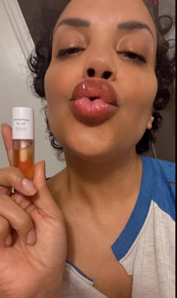 Person holding a lip oil, puckering lips for a selfie. Casual shirt, hair tied back. Suitable for beauty enthusiasts interested in lip care products