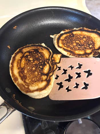 Two pancakes in a skillet, one being lifted with a spatula featuring bird cutouts
