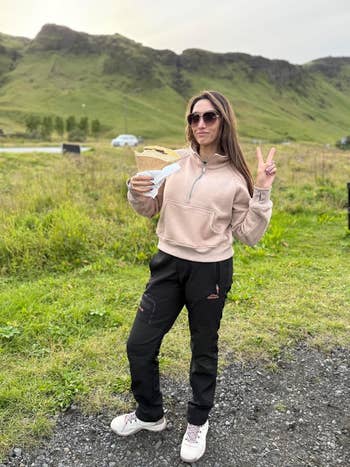 Person outdoors wearing casual pants and hoodie, holding a large sandwich and making a peace sign, with a scenic hillside background