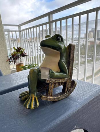 Frog figurine on a rocking chair holding a mug, placed on a balcony. Ideal for home or garden decor