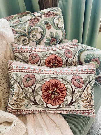Two embroidered throw pillows with floral patterns on a patterned armchair, draped with a soft, light-colored blanket