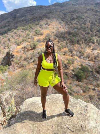 Person wearing a stylish activewear set and sunglasses, standing confidently on a rocky terrain with a scenic mountainous background
