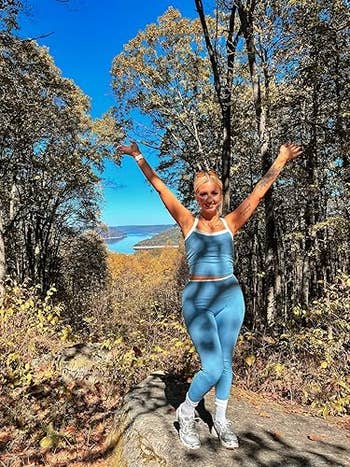 Person in athletic wear stands joyfully on a wooded trail, arms raised, with a lake and mountains visible in the background