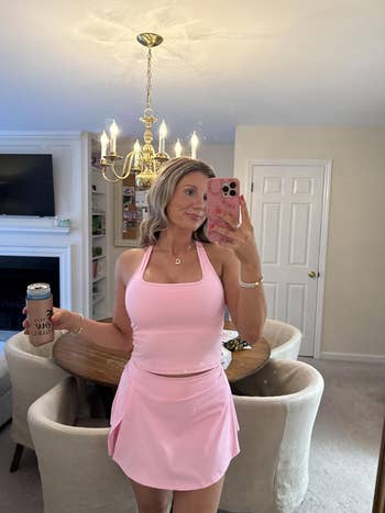 Person taking a mirror selfie in a sleeveless top and skirt, holding a can. Living room with a chandelier is visible in the background