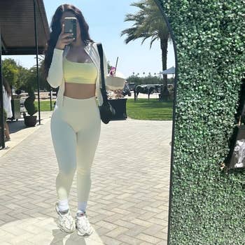 Person in activewear takes a mirror selfie outdoors. They're wearing a sports bra and leggings, with sneakers and a shoulder bag visible