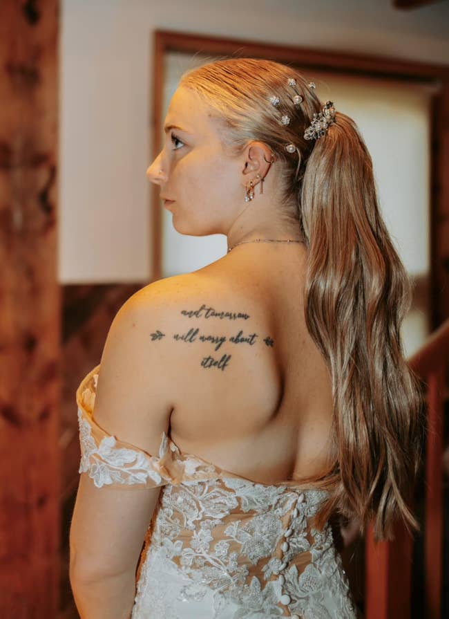 A reviewer in lace off-shoulder dress with long hair in a ponytail, adorned with small floral accessories, shows a tattoo on their upper back