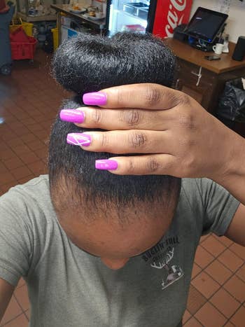 A reviewer with a high bun showcasing long, glossy purple nails with a geometric design, photographed in a casual indoor setting