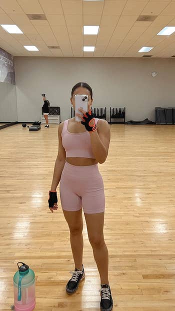 Person in a gym mirror selfie wearing activewear, fingerless gloves, and sneakers, with gym equipment visible in the background