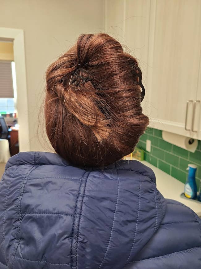 A reviewer with hair in a bun, wearing a quilted jacket. They're indoors, near kitchen cabinets and a green-tiled wall