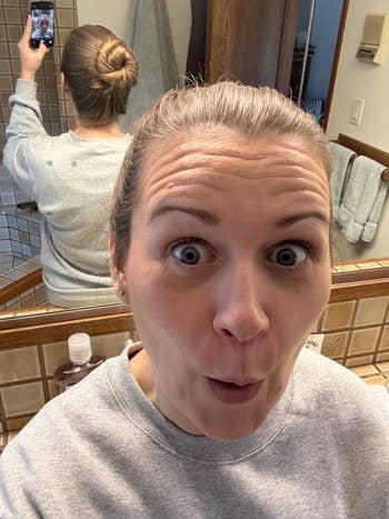 A reviewer with an expressive face takes a bathroom mirror selfie showing a neatly styled bun at the back of their head