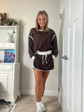Person smiling in a casual long-sleeve top and shorts set with sneakers, standing indoors
