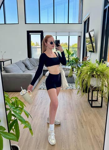 Person in a modern living room wears a stylish athletic outfit with sunglasses, holding a tumbler and posing for a mirror selfie
