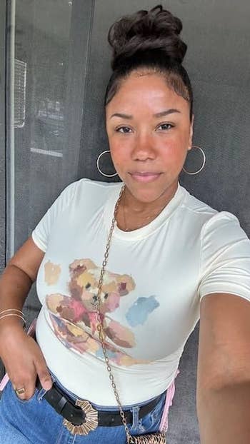 a reviewer wearing a printed t-shirt with a teddy bear design, gold hoop earrings, and blue jeans. Hair is styled in a bun