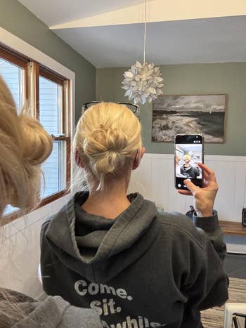 A reviewer with a bun hairstyle takes a mirror selfie, wearing a hooded sweatshirt that reads 